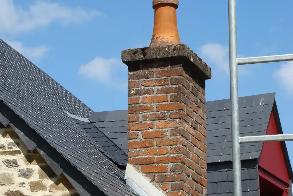 Cheminée en briques sur une toiture en ardoise, avec un échafaudage visible sous un ciel dégagé.