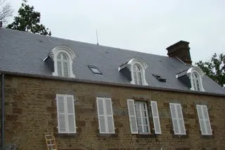 Façade d'une demeure ancienne en pierre, coiffée d'une toiture en ardoises avec plusieurs lucarnes.