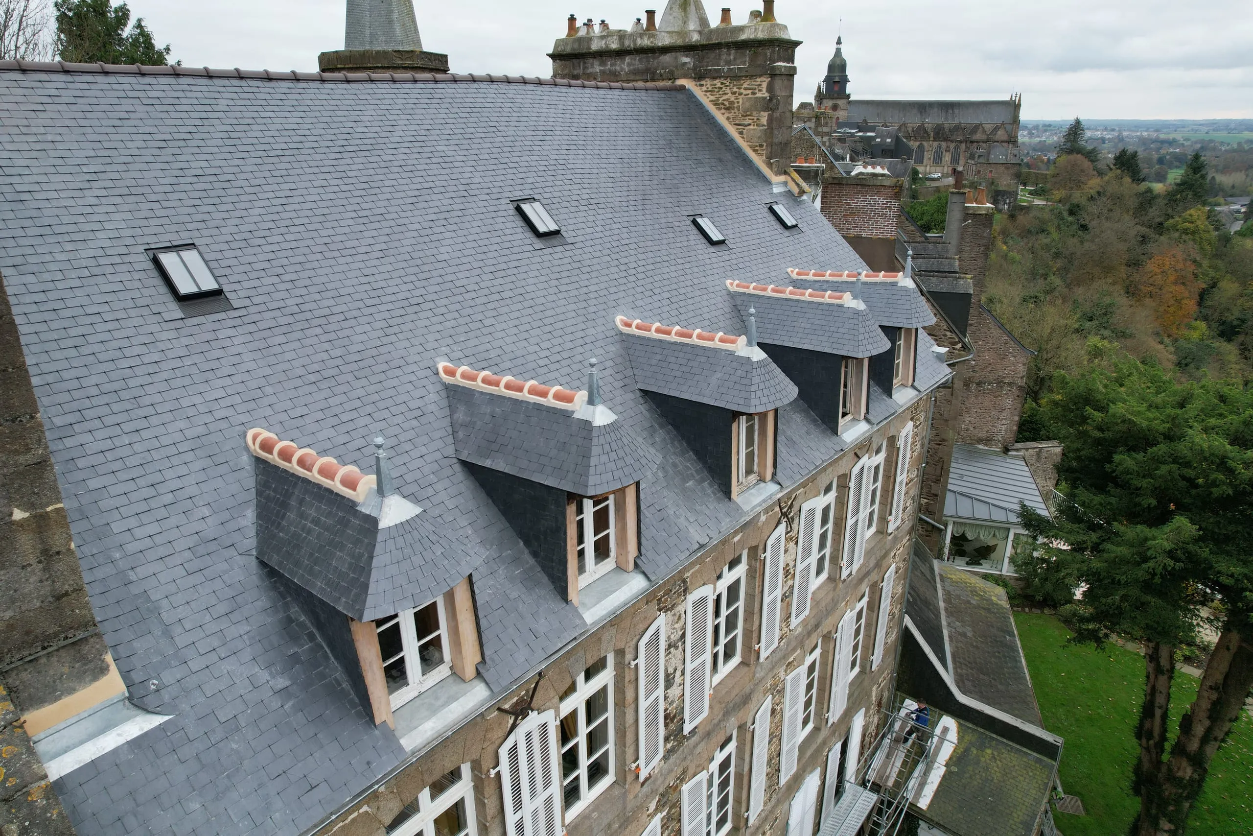 Vue aérienne d'une toiture en ardoise avec lucarnes à épis en zinc et fenêtres de toit sur un bâtiment en pierre